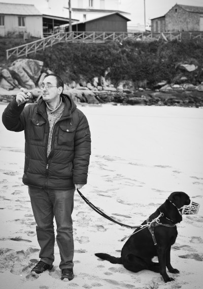Foto en branco e negro no que se pode ver a un home na praia facendo unha foto xunto co seu can guía