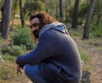 Mohamad Bitari agachado nun bosque, cunha expresión tranquila e sorrinte. Viste roupa informal con xersei azul e capucha gris. Ao fondo, árbores e vexetación nun espazo natural con luz suave de atardecer.