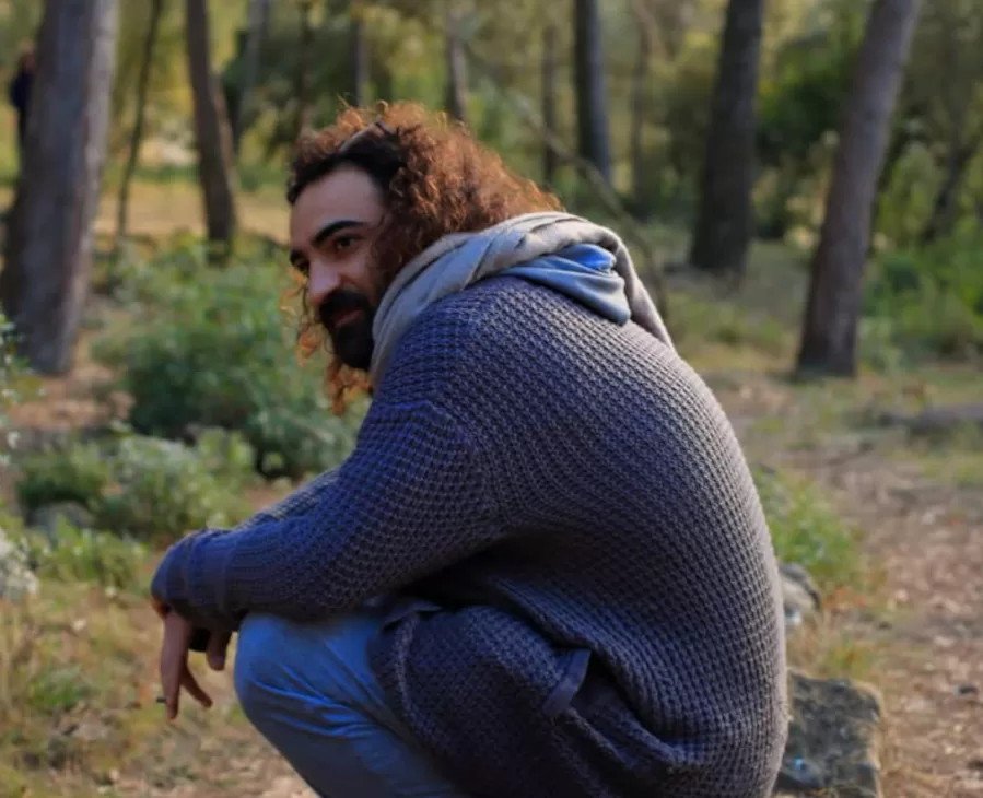 Mohamad Bitari agachado nun bosque, cunha expresión tranquila e sorrinte. Viste roupa informal con xersei azul e capucha gris. Ao fondo, árbores e vexetación nun espazo natural con luz suave de atardecer.
