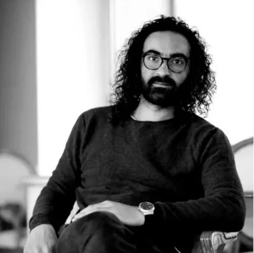 escrita no remoto 2025 Retrato en branco e negro de Mohamad Bitari sentado nunha cadeira, mirando á cámara. Leva lentes, ten o cabelo rizado e solto, e viste un xersei escuro. A expresión do rostro é seria e reflexiva