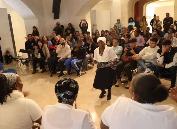 txt batukeiradas Un grupo de mulleres, as batukadeiras de Nos Eransa, durante unha actuación enérxica nun espazo interior. Cinco delas están sentadas en fila, aplaudindo e cantando con entusiasmo, mentres unha muller de máis idade, de pé cun sombreiro branco, baila no centro cun pequeno tambor na man.