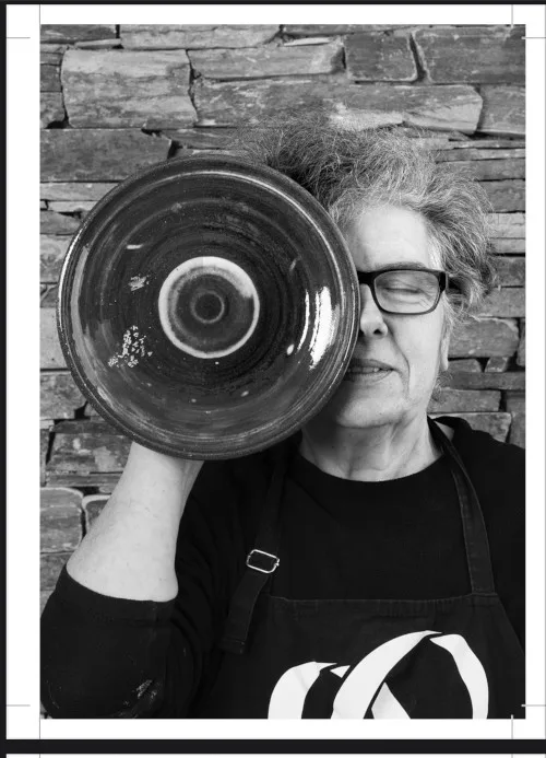 Retrato de plano medio en branco e negro dunha persoa con gafas e pelo ondulado, vestida cun mandil de traballo. A persoa suxeita unha cunca grande de cerámica esmaltada diante do seu rostro, tapando o ollo dereito, e mira á cámara co ollo esquerdo. O fondo está composto por unha parede de pedra.
