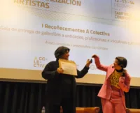 Lúa Gándara, vestida cun traxe rosa, entregando un premio (un pequeno obxecto azul) a Encarna Lago, Xerente da RMPL, nun escenario. Encarna Lago (á esquerda, de negro) sostén un diploma. Ao fondo, unha pantalla proxecta información sobre os "I Recoñecementos A Colectiva" e a gala de entrega de galardóns.