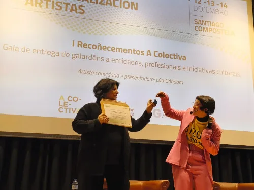 Lúa Gándara, vestida cun traxe rosa, entregando un premio (un pequeno obxecto azul) a Encarna Lago, Xerente da RMPL, nun escenario. Encarna Lago (á esquerda, de negro) sostén un diploma. Ao fondo, unha pantalla proxecta información sobre os "I Recoñecementos A Colectiva" e a gala de entrega de galardóns.