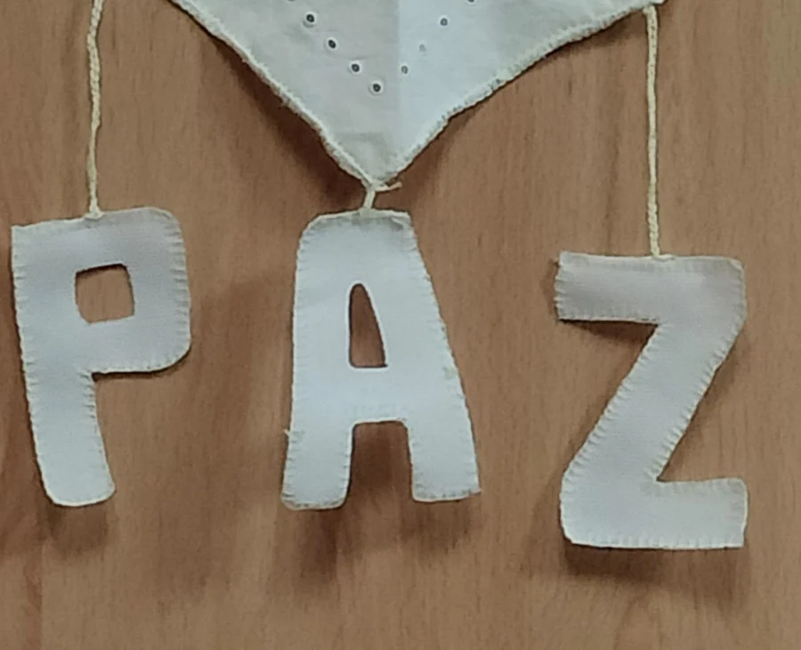 Pezas de tea branca penduradas nunha parede formando a composición "Fíos de Paz". Na parte superior, un pano bordado sostén as figuras de dúas pombas brancas que suxeitan unha póla de oliveira. Da parte inferior do pano colgan tres letras de tea que forman a palabra "PAZ".