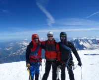 Tres montañeiros do equipo de 3Cumios equipados con roupa técnica, cordas e cascos, posan nun cume nevado dos Andes baixo un ceo despexado, coa cordilleira estendéndose ao fondo.