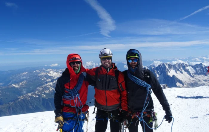 Tres montañeiros do equipo de 3Cumios equipados con roupa técnica, cordas e cascos, posan nun cume nevado dos Andes baixo un ceo despexado, coa cordilleira estendéndose ao fondo.