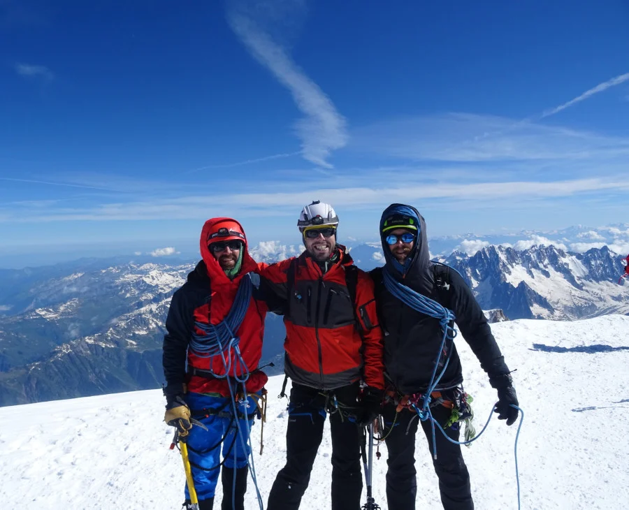 Tres montañeiros do equipo de 3Cumios equipados con roupa técnica, cordas e cascos, posan nun cume nevado dos Andes baixo un ceo despexado, coa cordilleira estendéndose ao fondo.