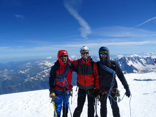 Tres montañeiros do equipo de 3Cumios equipados con roupa técnica, cordas e cascos, posan nun cume nevado dos Andes baixo un ceo despexado, coa cordilleira estendéndose ao fondo.